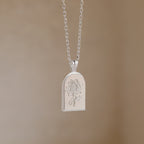 The Birth Flower Arch Necklace features an engraved floral design in silver, displayed against a neutral beige background—a stunning piece of personalized jewelry.
