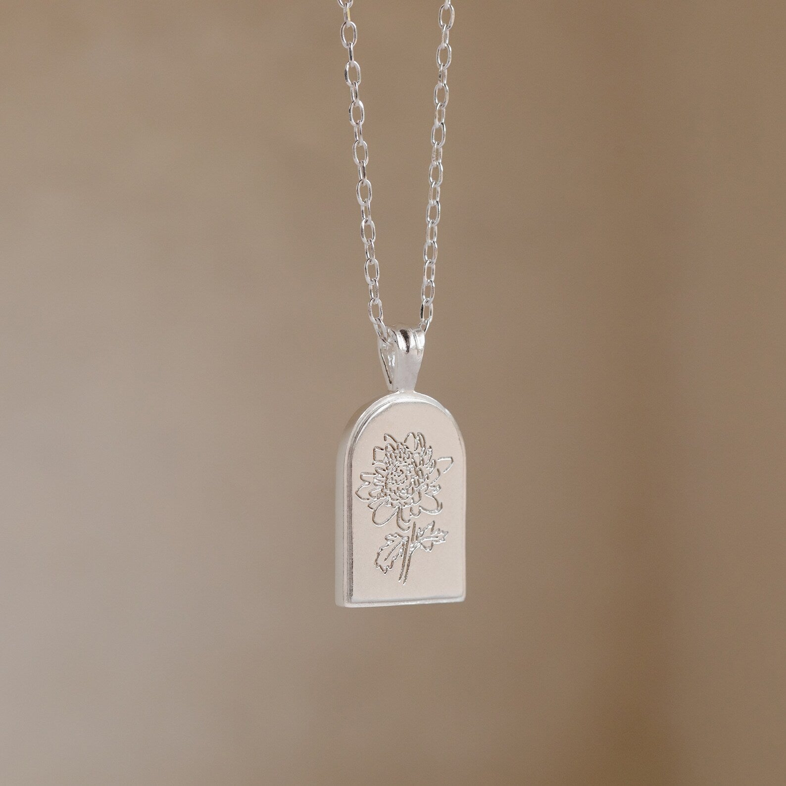 The Birth Flower Arch Necklace features an engraved floral design in silver, displayed against a neutral beige background—a stunning piece of personalized jewelry.