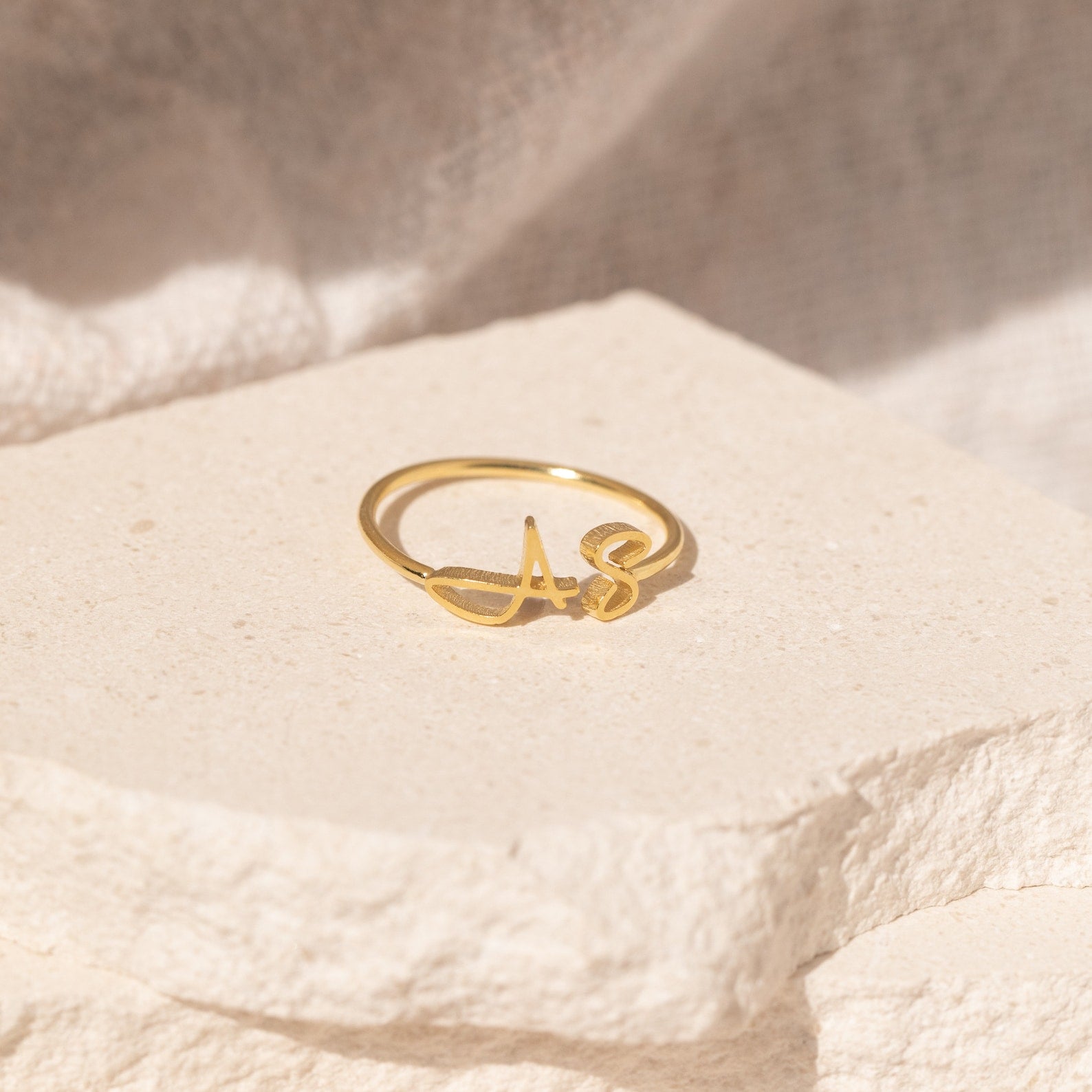 The Hallie Duo Initial Ring, featuring "AS," rests on a textured, light-colored stone surface—a stylish personalized jewelry piece and an ideal gift for moms.