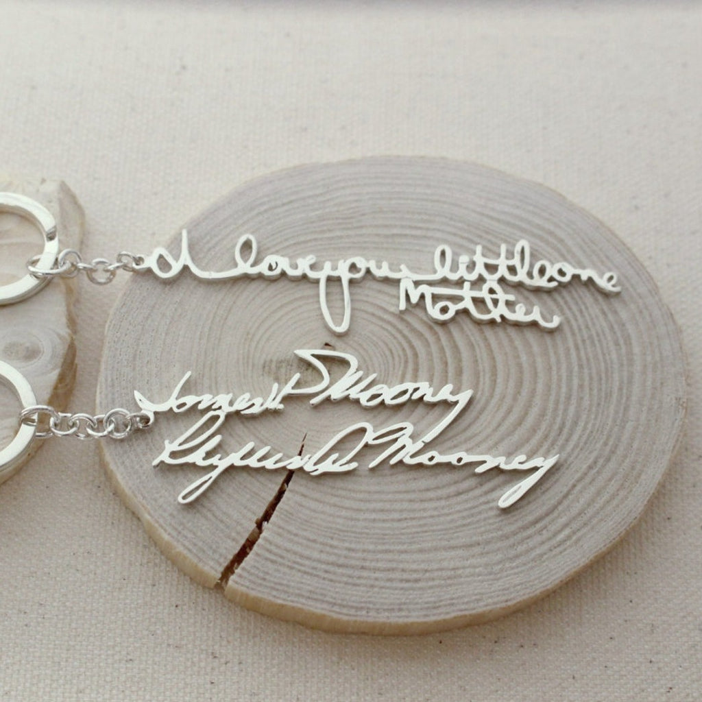 Showcasing two silver necklaces with cursive handwriting pendants on wood; one says "I love you little one Mother." These unique pieces pair perfectly with our Custom Handwriting Keychain—ideal for heartfelt, personalized gifts.