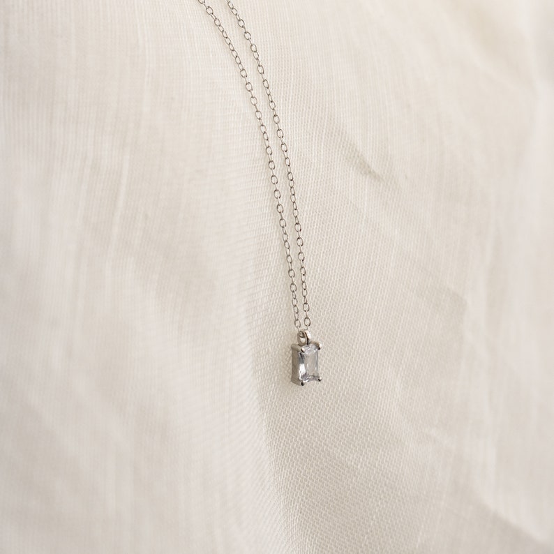 The Sage Diamond Necklace features a delicate silver chain and a small rectangular pendant, reflecting timeless elegance against a soft white fabric background.