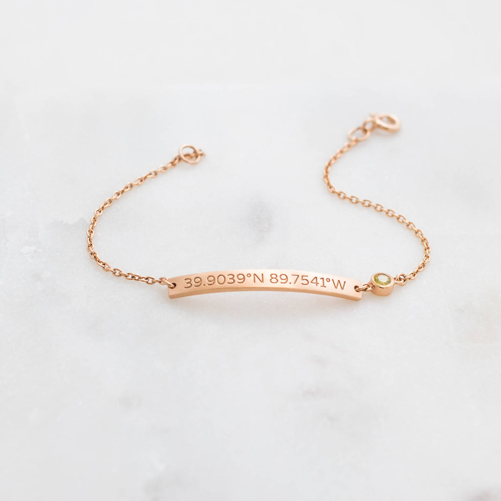 The Thin Coordinates Bar Bracelet in rose gold, engraved with 39.9039°N 89.7541°W, rests on a white surface—ideal personalized jewelry to commemorate a special location.