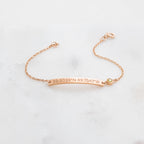 The Thin Coordinates Bar Bracelet in rose gold, engraved with 39.9039°N 89.7541°W, rests on a white surface—ideal personalized jewelry to commemorate a special location.