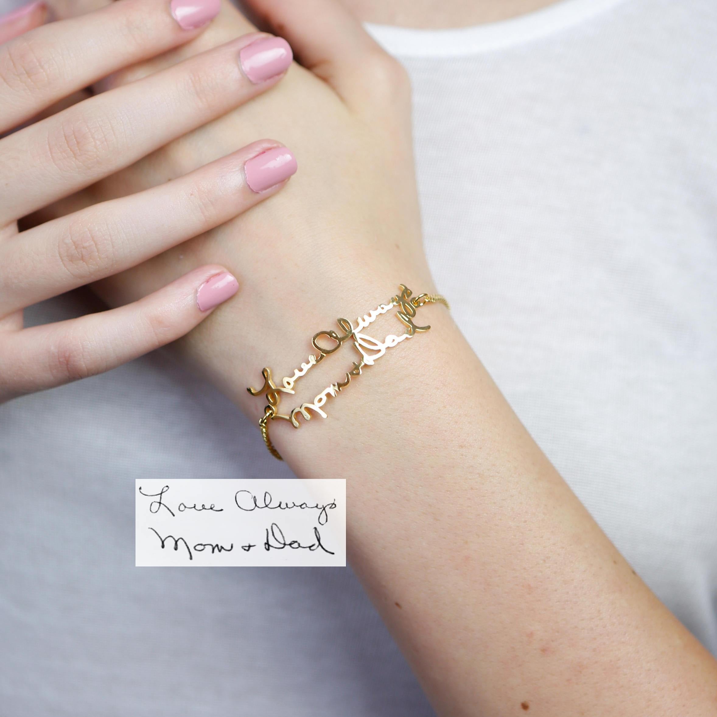 A Handwriting Bracelet in gold, featuring "Love Always Mom & Dad" in cursive, worn with light pink nails—ideal for daily wear or as a meaningful piece of memorial jewelry.