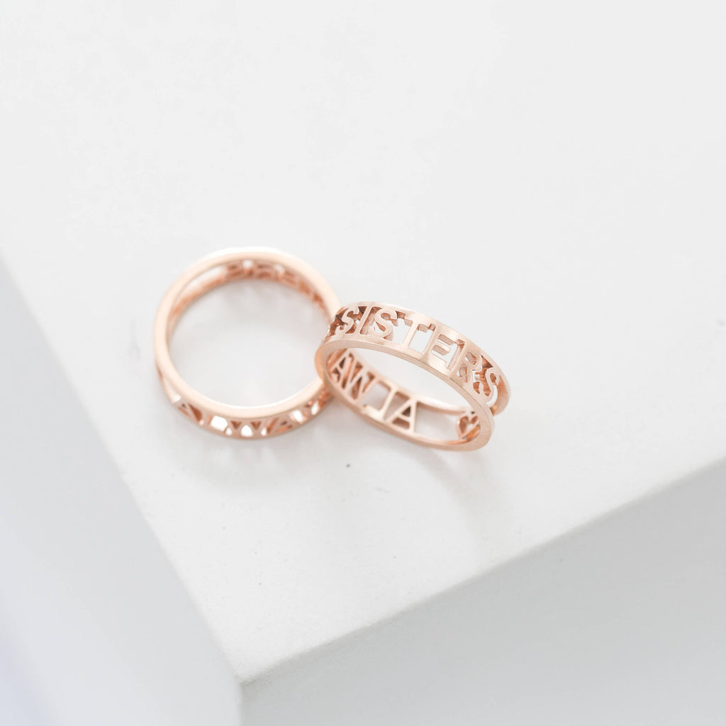 Two rose gold rings rest on a white surface—one engraved with SISTERS, the other is the Roman Numerals Ring in Rose Gold, ideal for commemorating a special date or personal milestone.