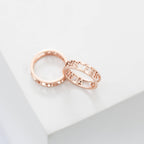 Two rose gold rings rest on a white surface—one engraved with SISTERS, the other is the Roman Numerals Ring in Rose Gold, ideal for commemorating a special date or personal milestone.