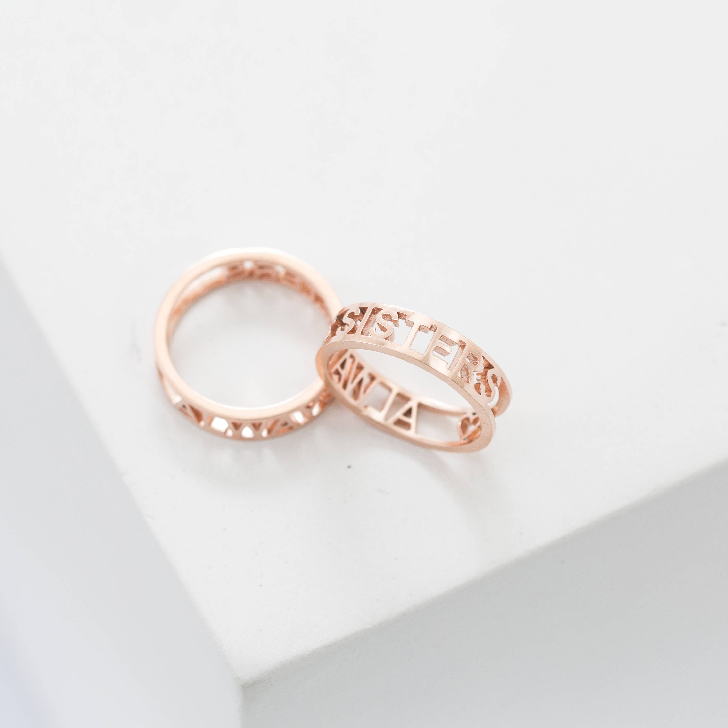 Two rose gold rings rest on a white surface—one engraved with SISTERS, the other is the Roman Numerals Ring in Rose Gold, ideal for commemorating a special date or personal milestone.