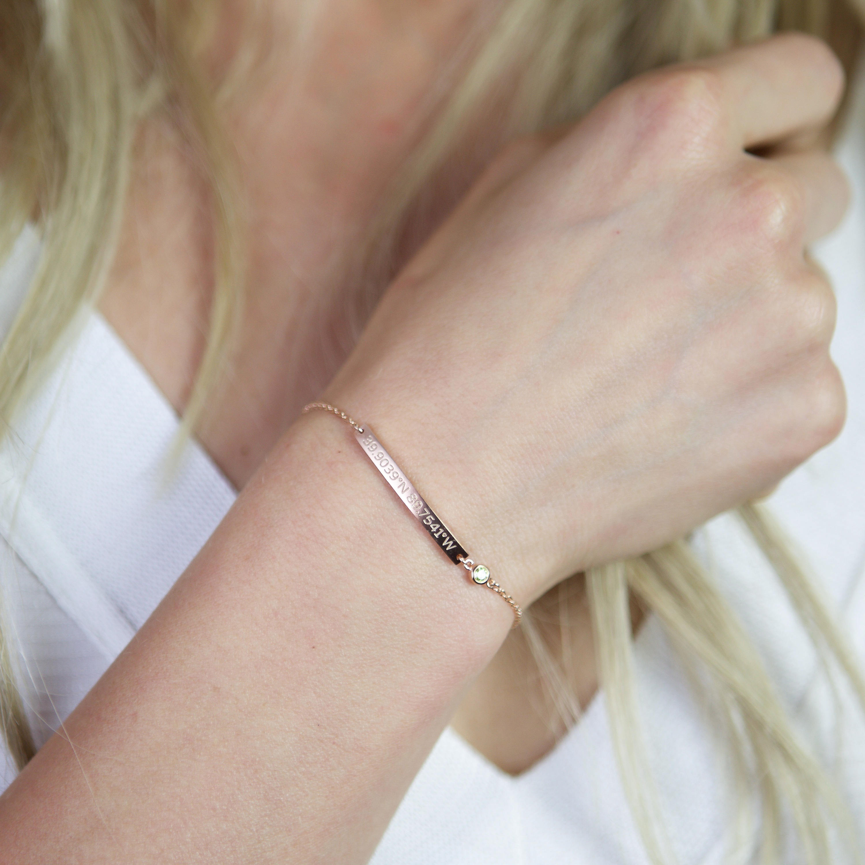A person wears a Thin Coordinates Bar Bracelet with a small gem on their wrist against a white top—ideal custom jewelry for location lovers.