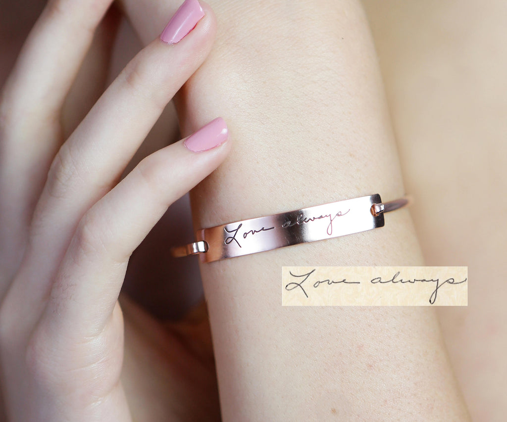 A hand with pink nails wears a Handwriting Bar Bangle engraved with "Love always" in cursive.