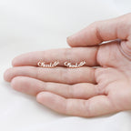 A hand holds two London Name Earrings shaped like the word "Charlotte" in elegant script font, perfect as personalized stud earrings.