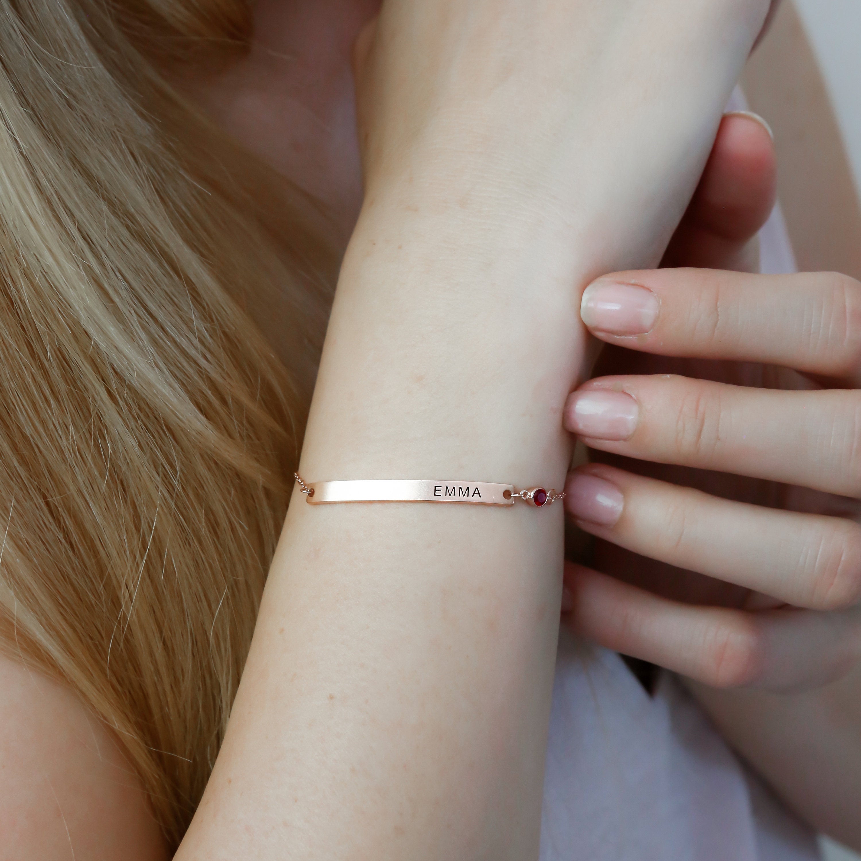 A woman wears a Thin Name Bar Bracelet engraved with EMMA on her wrist, showcasing elegant personalized jewelry.