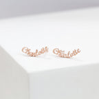 Two London Name Earrings rest on a white surface, each crafted in gold and elegantly shaped to spell "Charlotte" in a graceful script font.