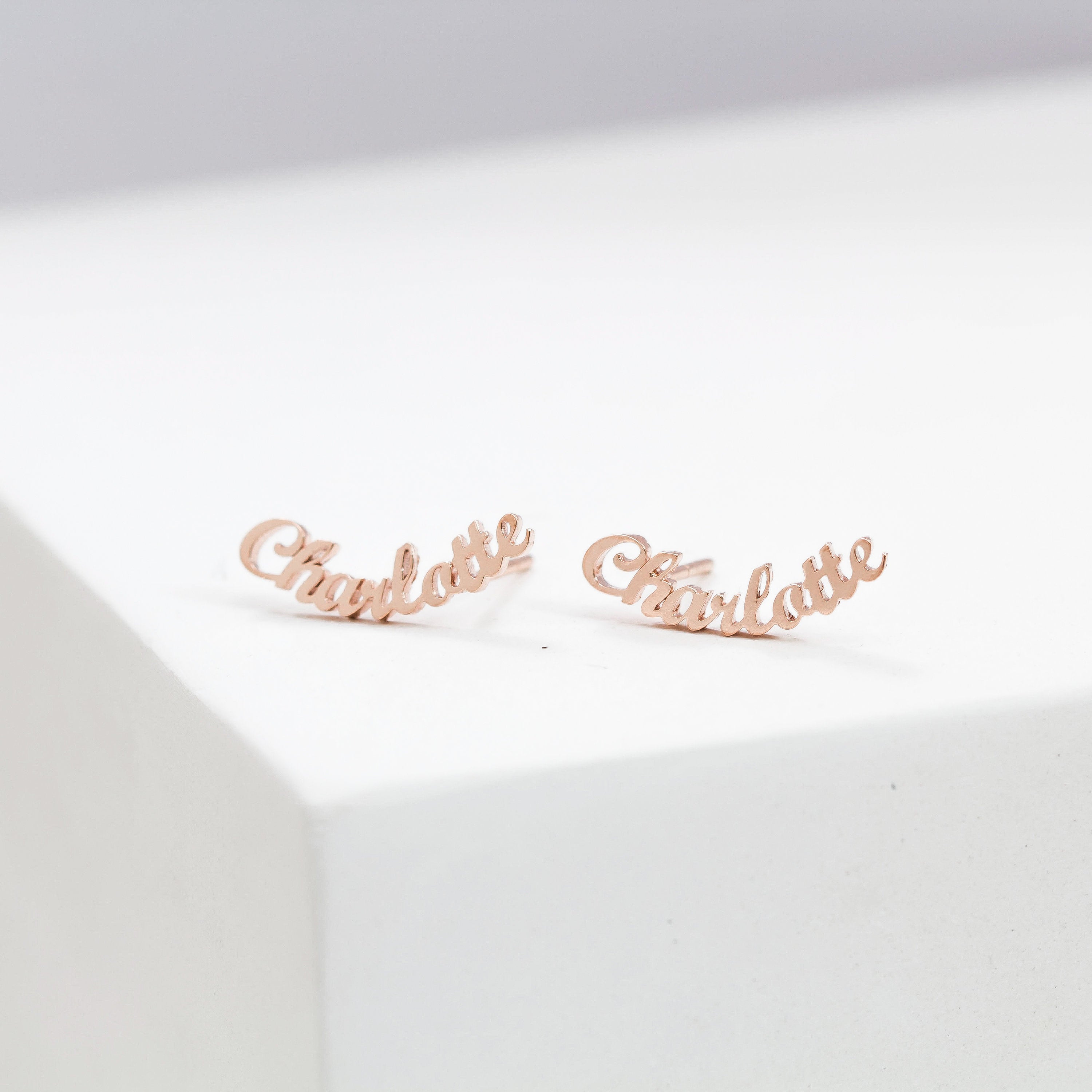 Two London Name Earrings rest on a white surface, each crafted in gold and elegantly shaped to spell "Charlotte" in a graceful script font.