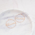 Two Classic Name Hoops, personalized with "Lauren," are beautifully displayed on a round white marble dish—ideal as standout custom jewelry.