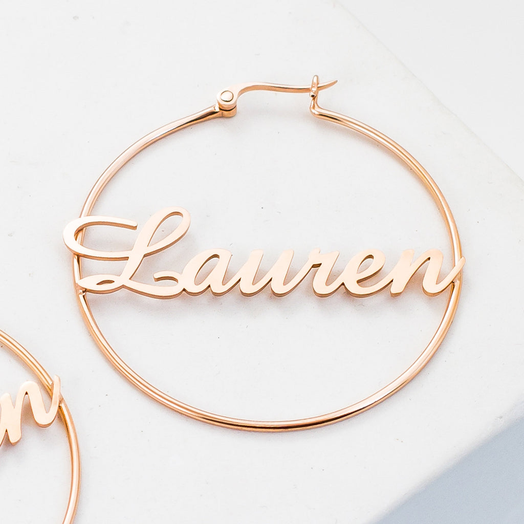 Classic Name Hoops feature gold hoop earrings with the name Lauren in cursive across the center—ideal for personalized jewelry lovers and stylish accessory fans, shown on a white surface.