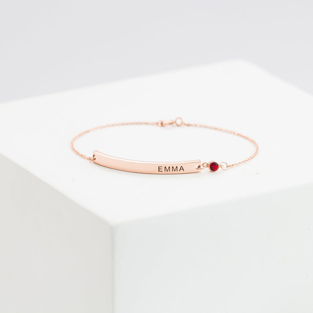 Thin Name Bar Bracelet in rose gold with personalized EMMA engraving and a small red gemstone, elegantly displayed on a white block.