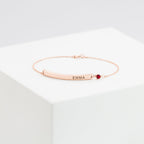Thin Name Bar Bracelet in rose gold with personalized EMMA engraving and a small red gemstone, elegantly displayed on a white block.