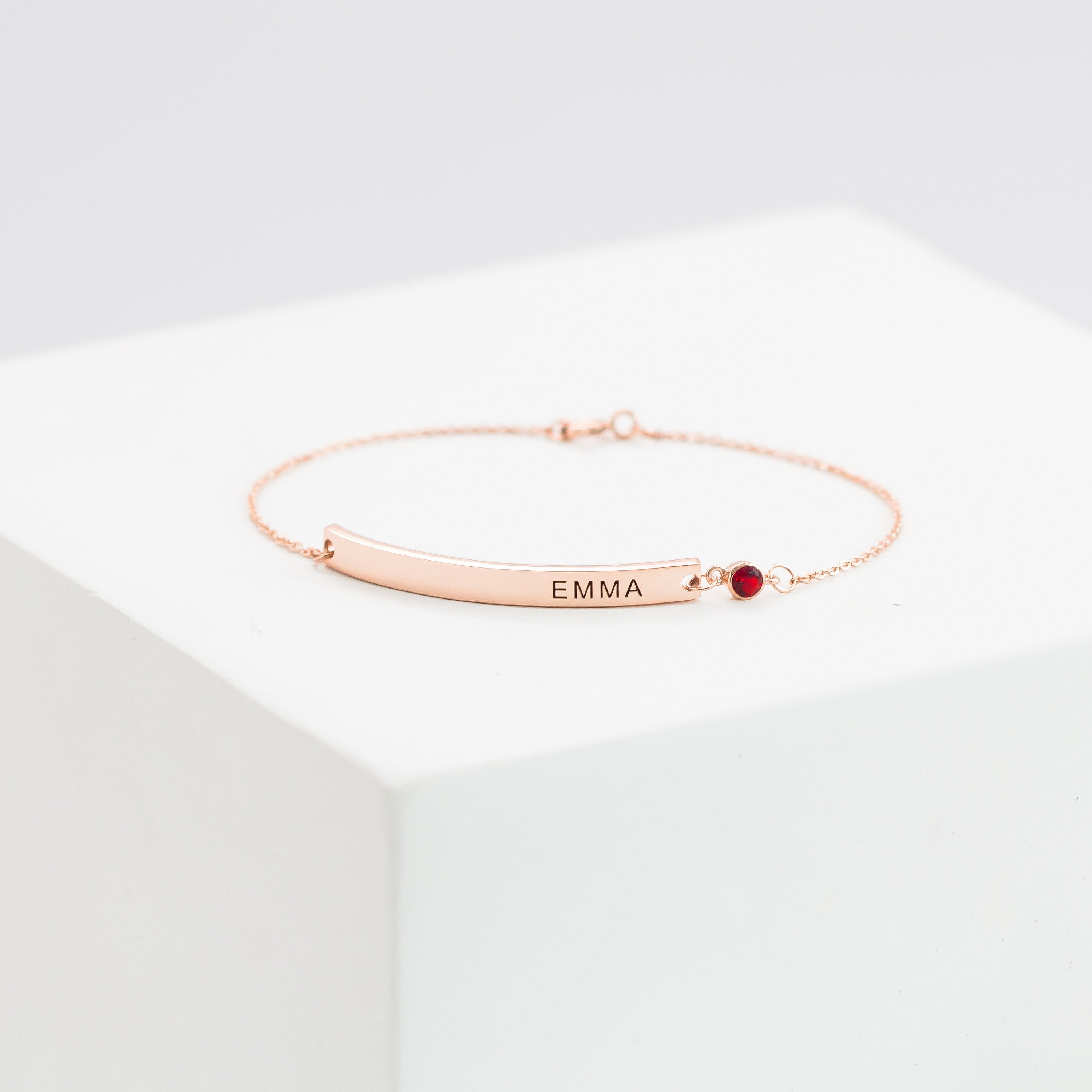 Thin Name Bar Bracelet in rose gold with personalized EMMA engraving and a small red gemstone, elegantly displayed on a white block.