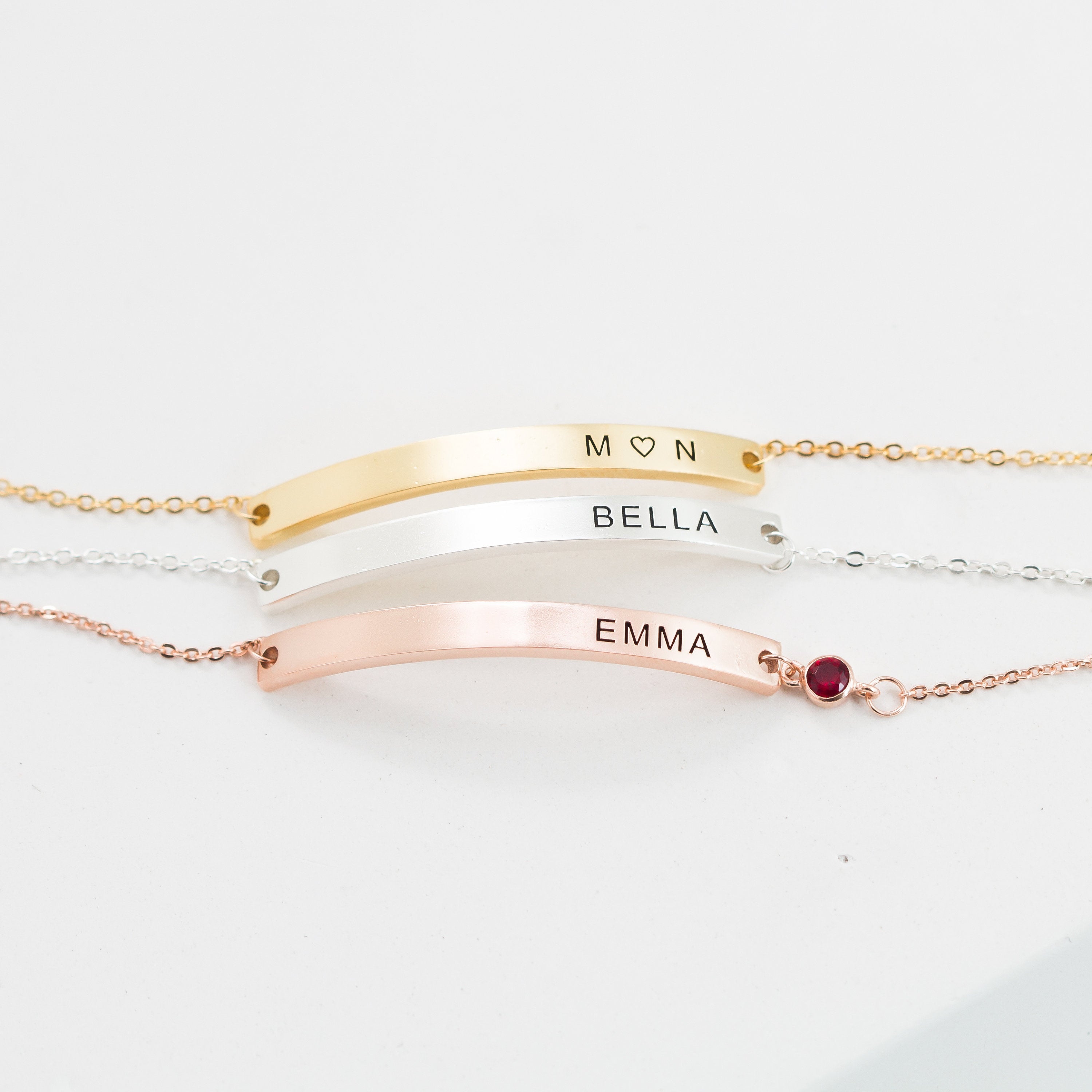 Three Thin Name Bar Bracelets in gold, silver, and rose gold—engraved M♡N, BELLA, and EMMA (with a red gem)—make stunning personalized jewelry for any dainty bracelet collection.