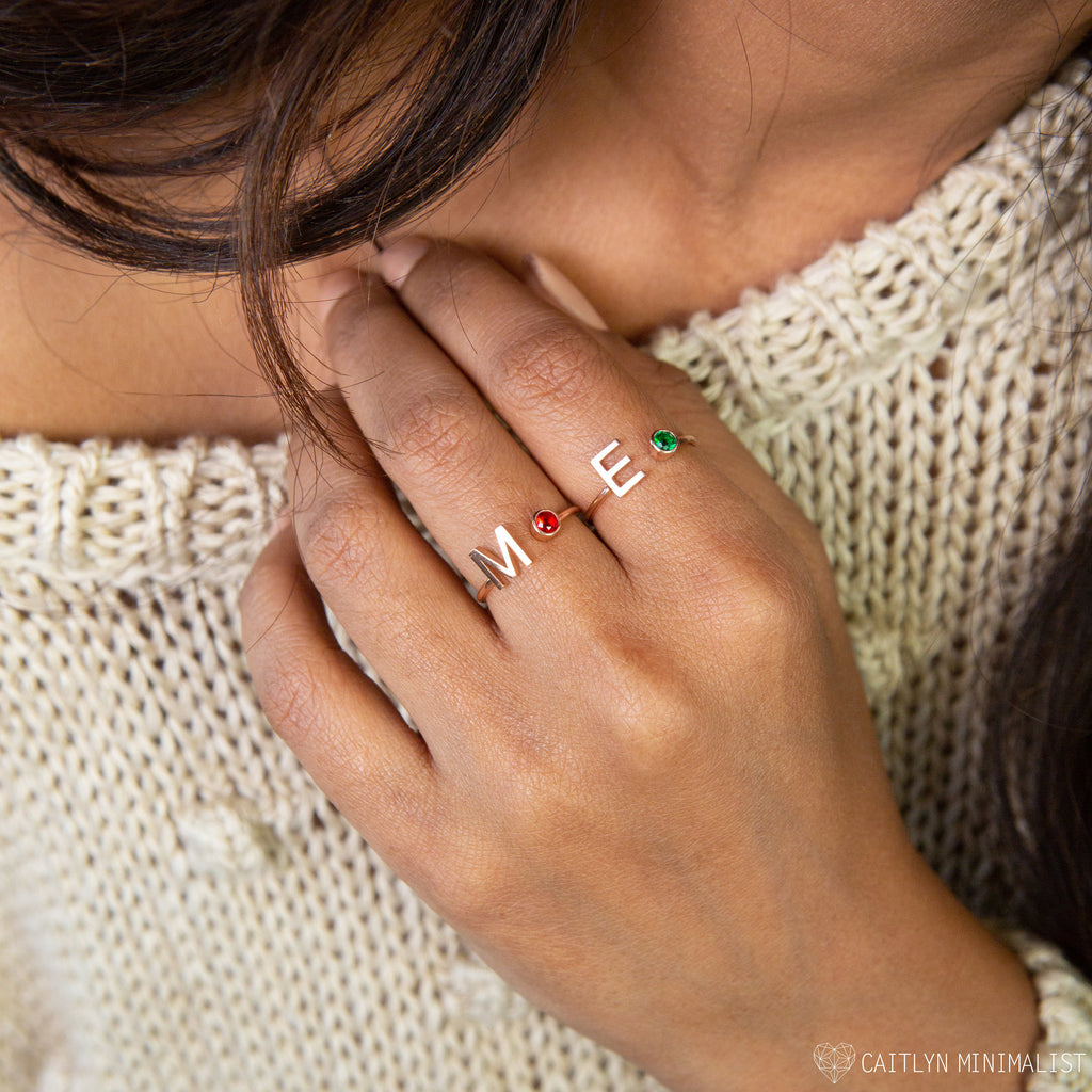 A hand with two Alex Initial Birthstone Rings in Rose Gold, each showcasing a red or green stone, on a cream knit sweater—perfect as personalized rings for a unique touch.