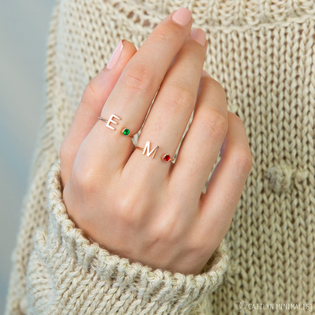 A hand wearing two Alex Initial Birthstone Rings—each with a colored gem and initial—paired with a cream knit sweater.