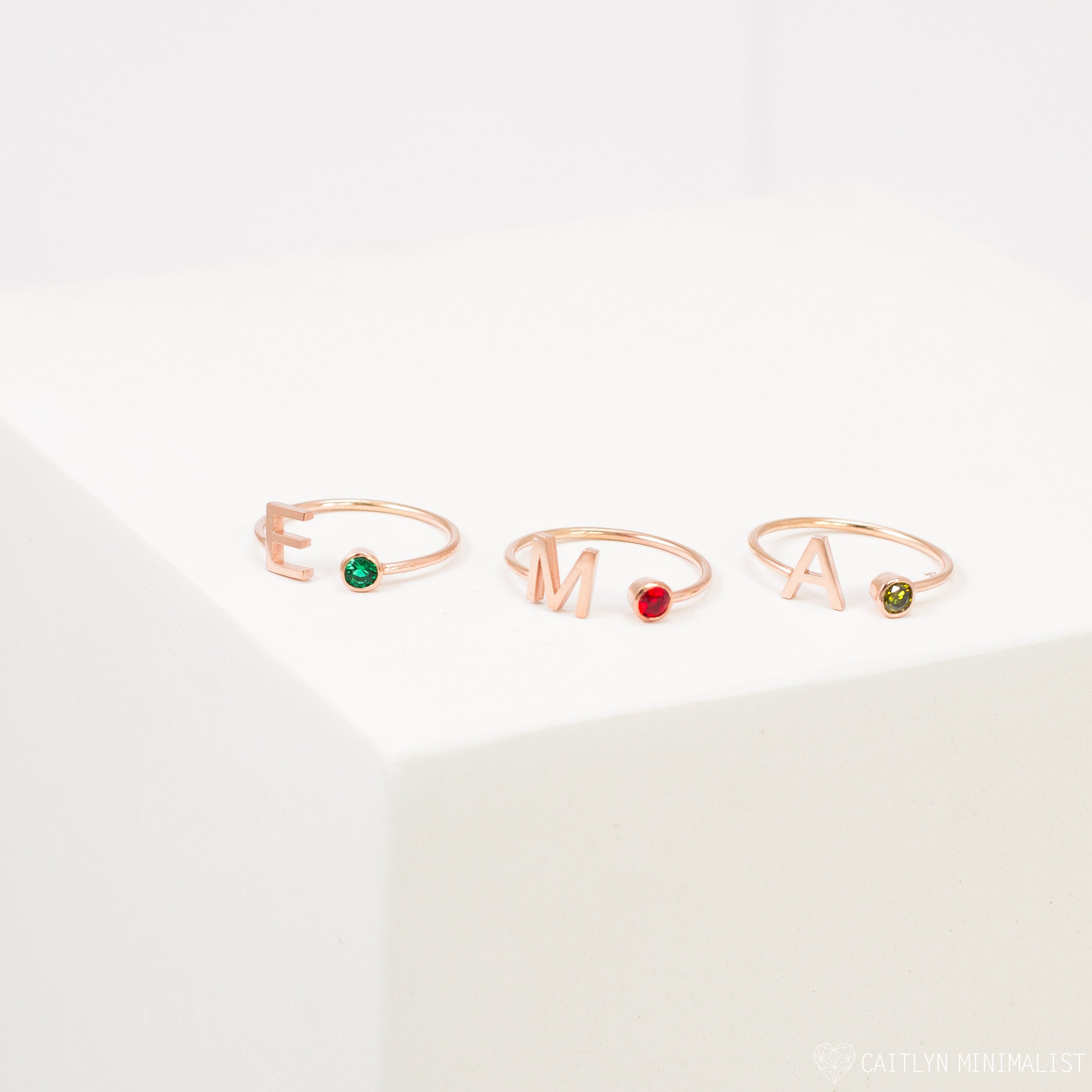 Three Alex Initial Birthstone Rings, each with letters E, M, and A and a colored gemstone, are displayed on a white surface—showcasing personalized wrap ring jewelry.