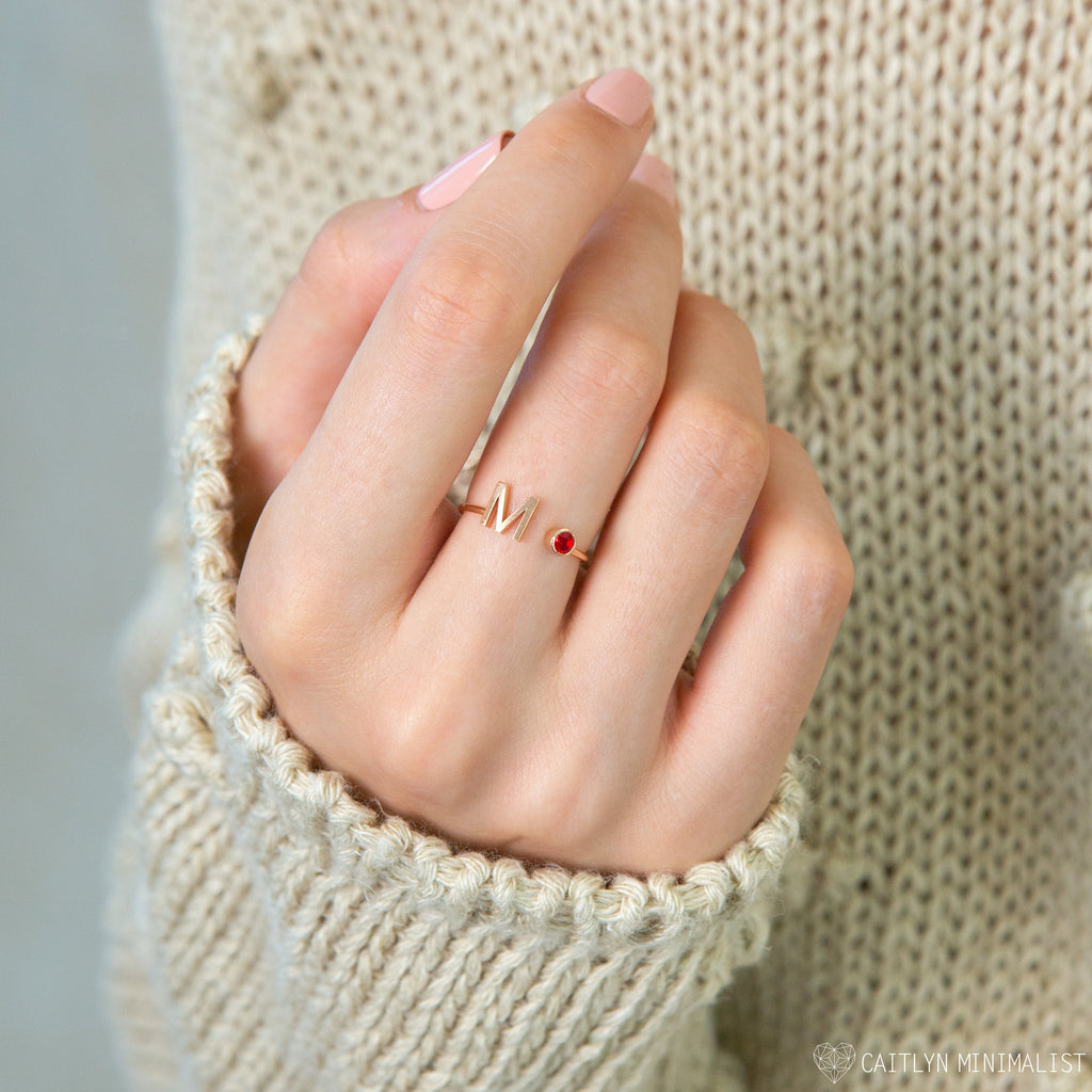 A hand with pink nails wears the Alex Initial Birthstone Ring, showcasing a gold M and red stone, against a beige knit sweater.