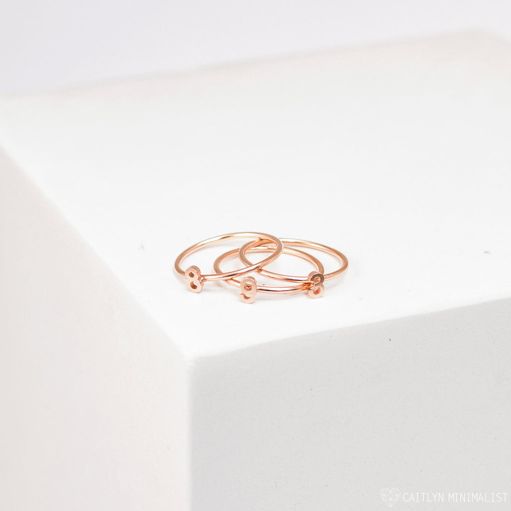 The Alex Dainty Letter Ring in Rose Gold, featuring delicate heart-shaped accents, is displayed alongside two matching rose gold rings on a white geometric surface.