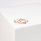 The Alex Dainty Letter Ring in Rose Gold, featuring delicate heart-shaped accents, is displayed alongside two matching rose gold rings on a white geometric surface.