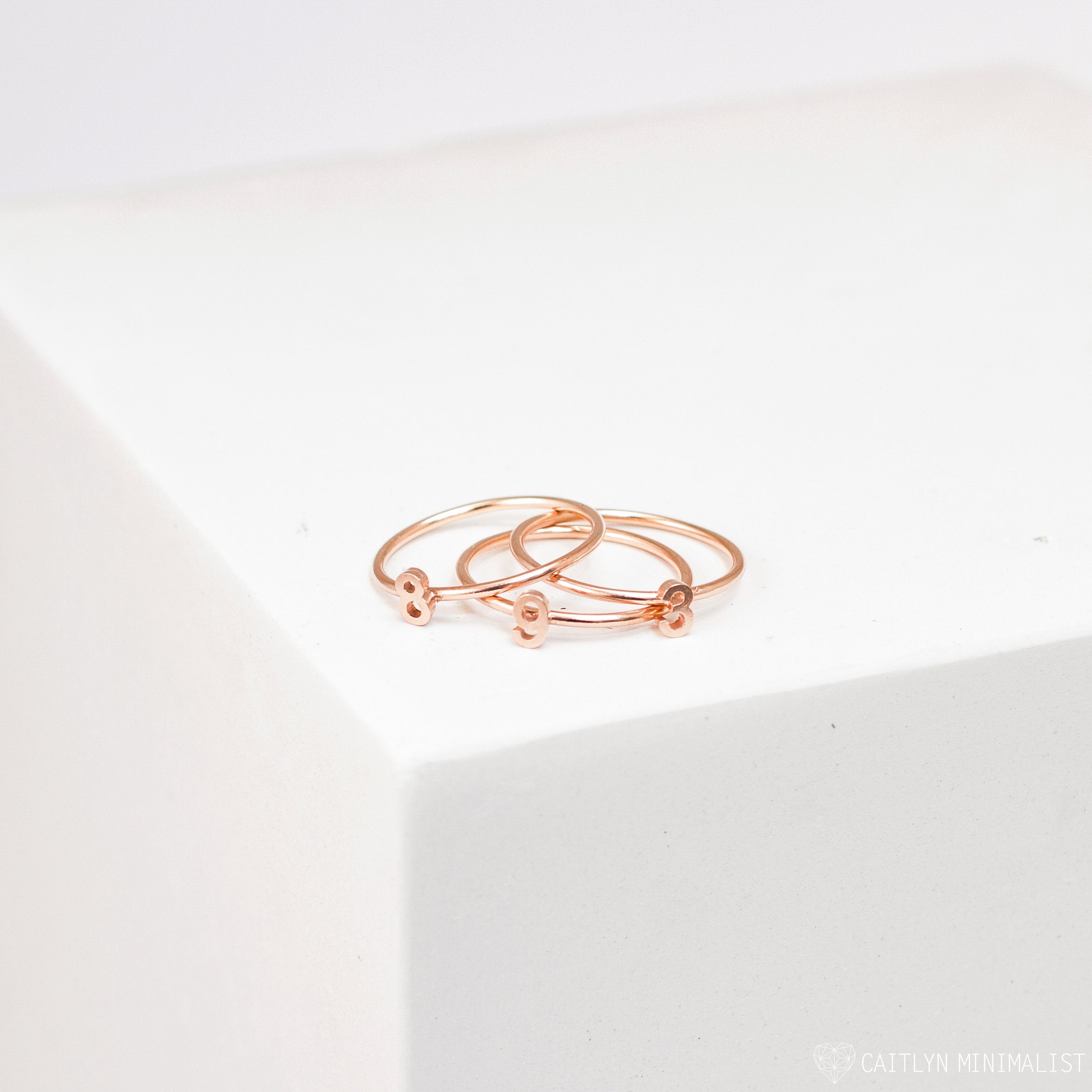 The Alex Dainty Letter Ring in Rose Gold, featuring delicate heart-shaped accents, is displayed alongside two matching rose gold rings on a white geometric surface.