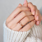 A hand with pale pink nails wears stacked gold rings, including the Alex Dainty Letter Ring in Rose Gold, shown against a white knit sweater background.