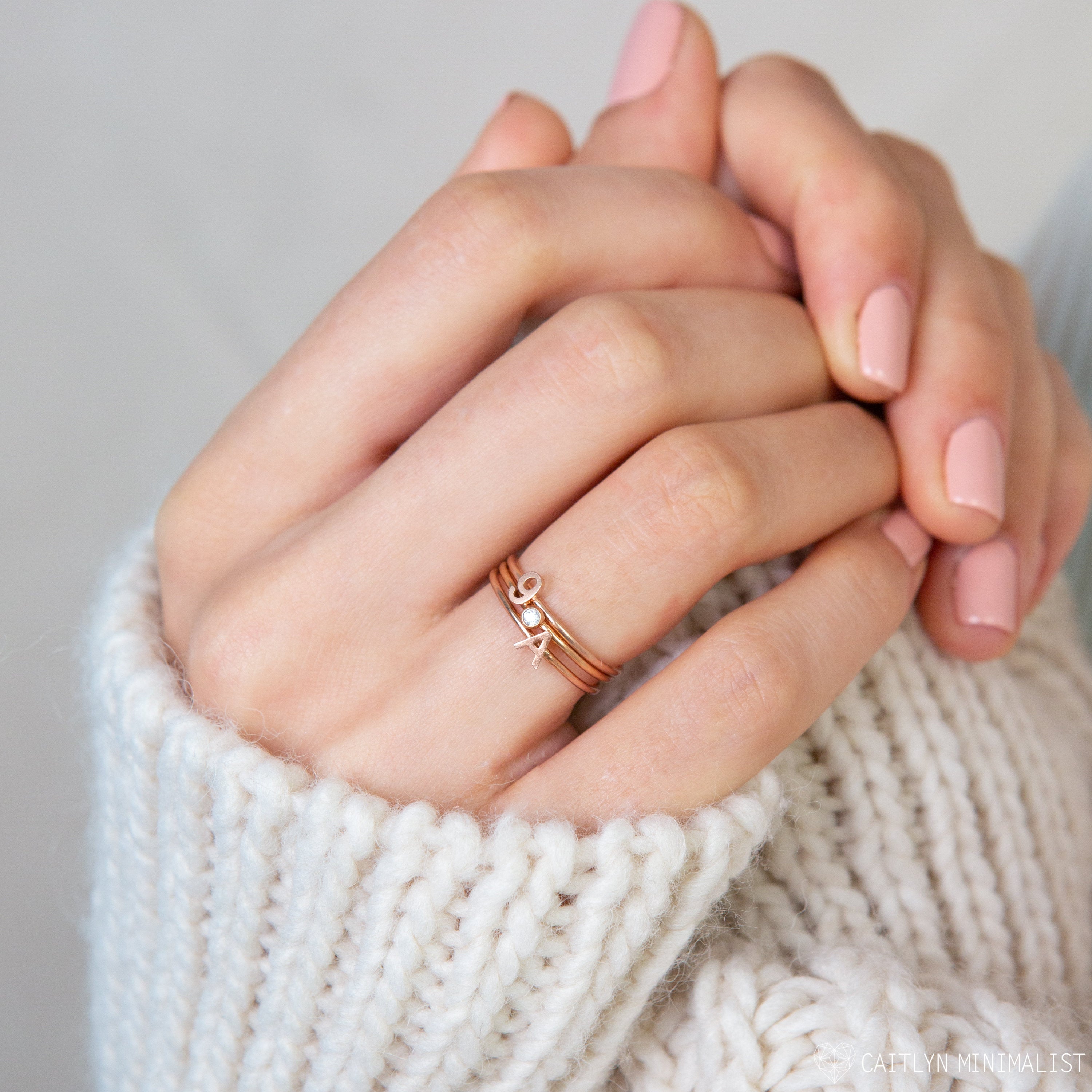 A hand with pale pink nails wears stacked gold rings, including the Alex Dainty Letter Ring in Rose Gold, shown against a white knit sweater background.