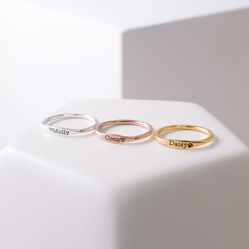 Three Arthur Skinny Name Rings in silver, rose gold, and gold—each engraved with Molly, Coco, and Daisy—are elegantly displayed on a white surface.