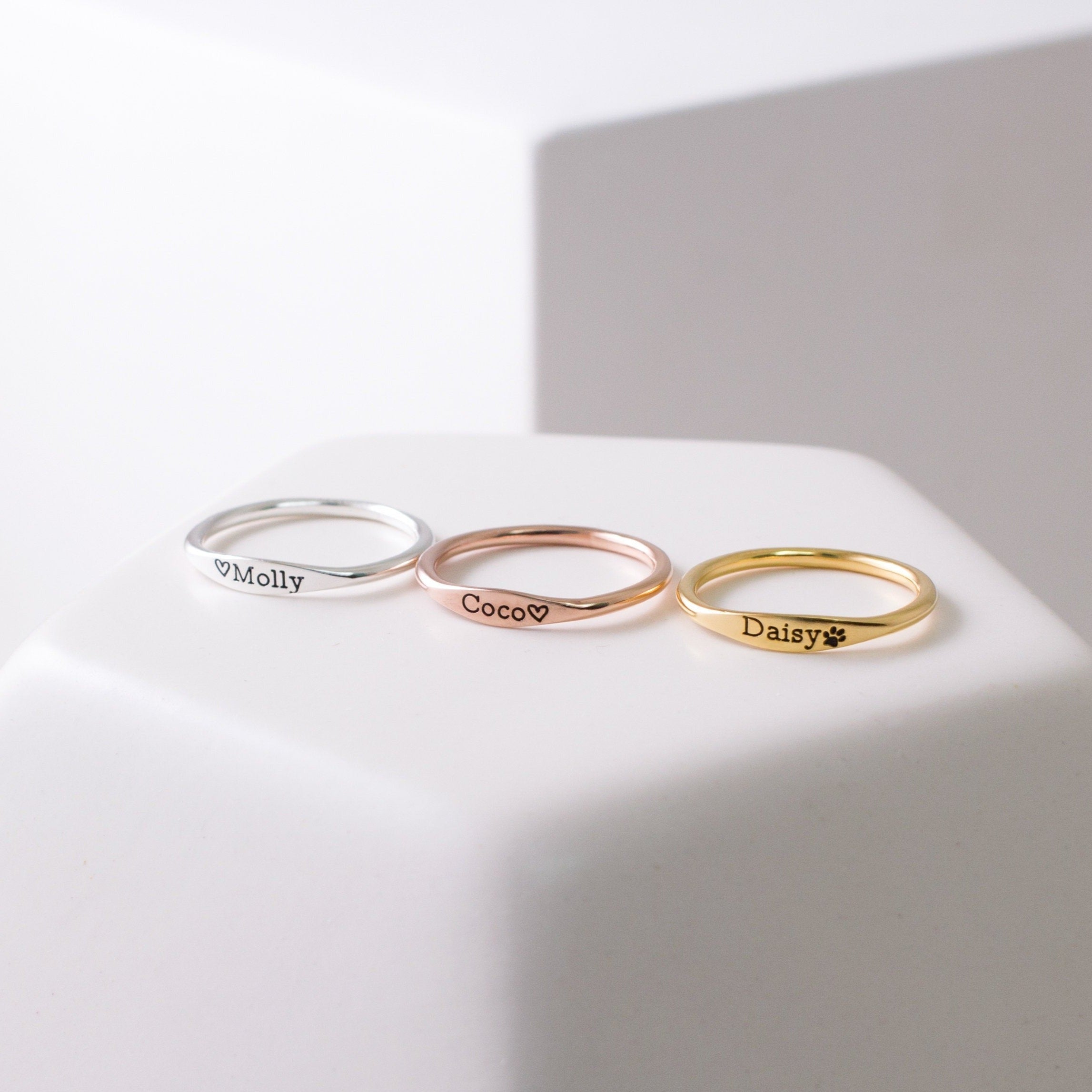 Three Arthur Skinny Name Rings in silver, rose gold, and gold—each engraved with Molly, Coco, and Daisy—are elegantly displayed on a white surface.