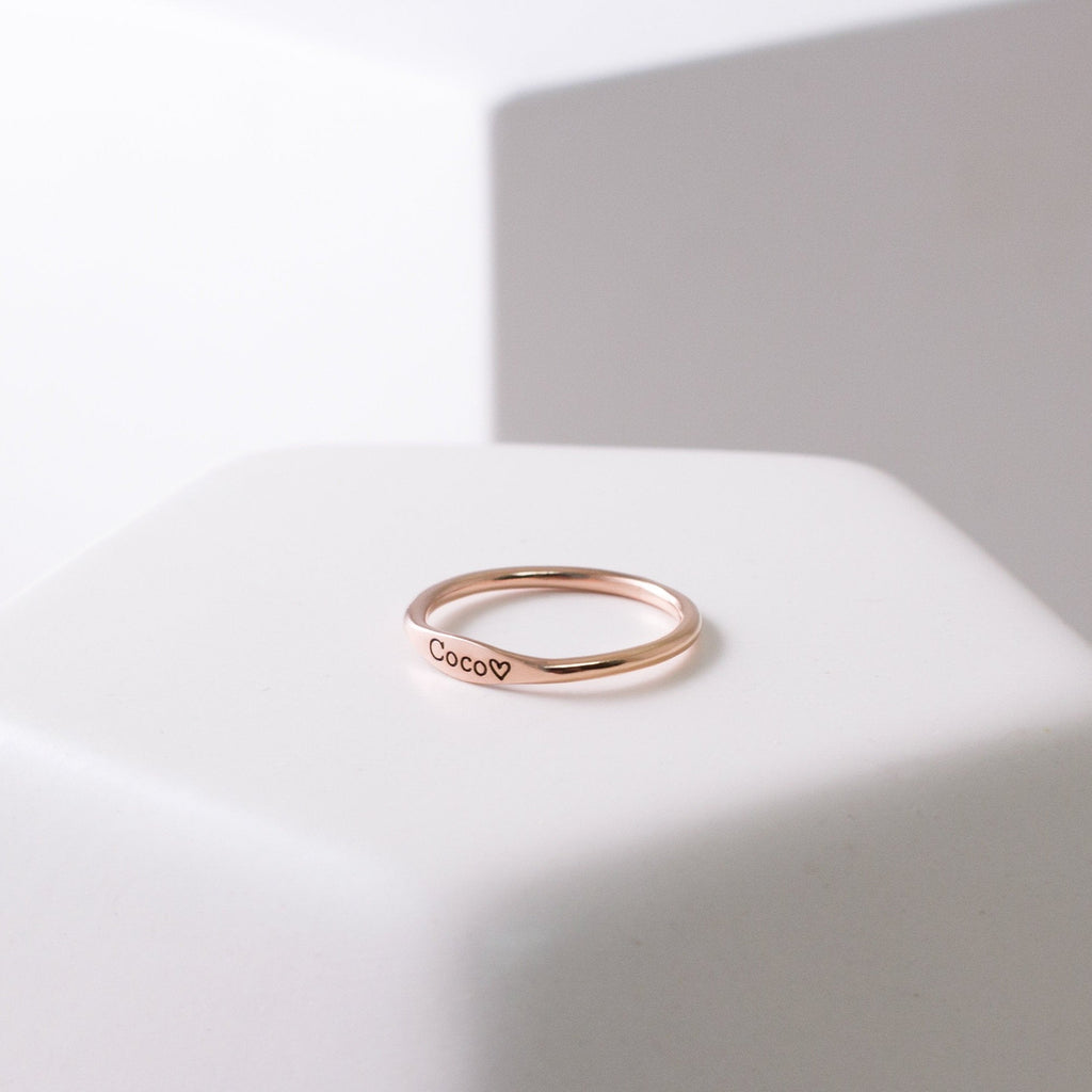 The Arthur Skinny Name Ring, a thin personalized ring engraved with Coco and a small heart, is displayed on a white surface.