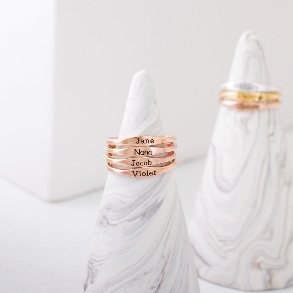 Four Arthur Skinny Name Rings in rose gold, each engraved with a name, are stacked on a marble cone holder; personalized skinny name rings and gold rings can be seen in the background.