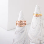 Four Arthur Skinny Name Rings in rose gold, each engraved with a name, are stacked on a marble cone holder; personalized skinny name rings and gold rings can be seen in the background.