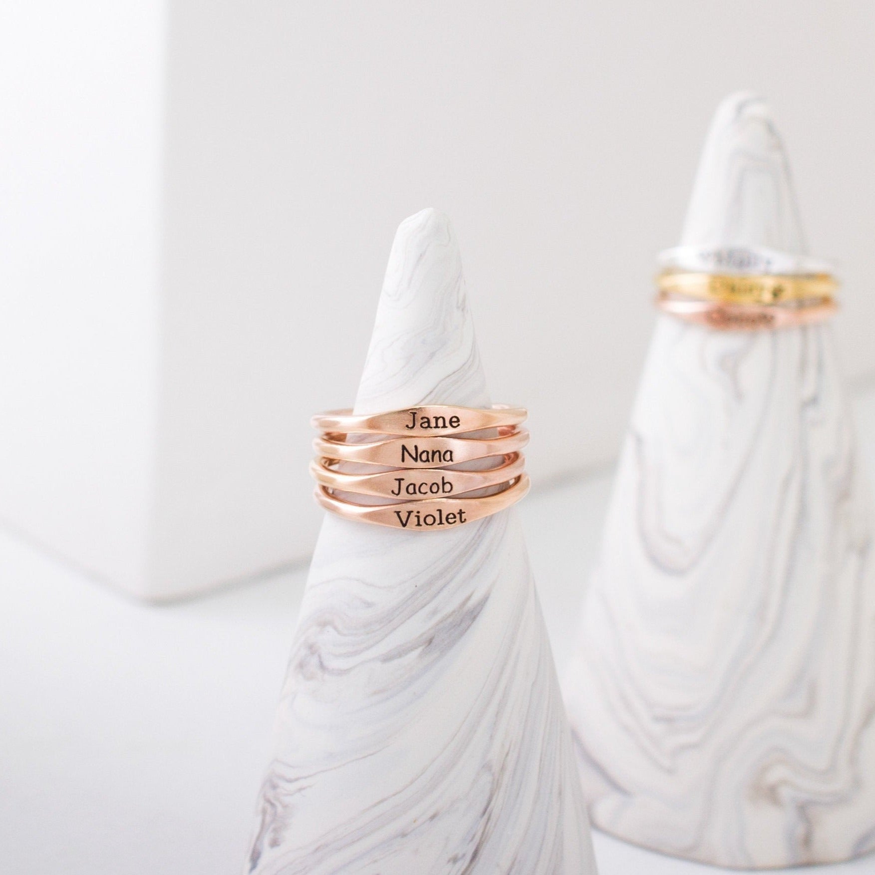 Four Arthur Skinny Name Rings in rose gold, each engraved with a name, are stacked on a marble cone holder; personalized skinny name rings and gold rings can be seen in the background.