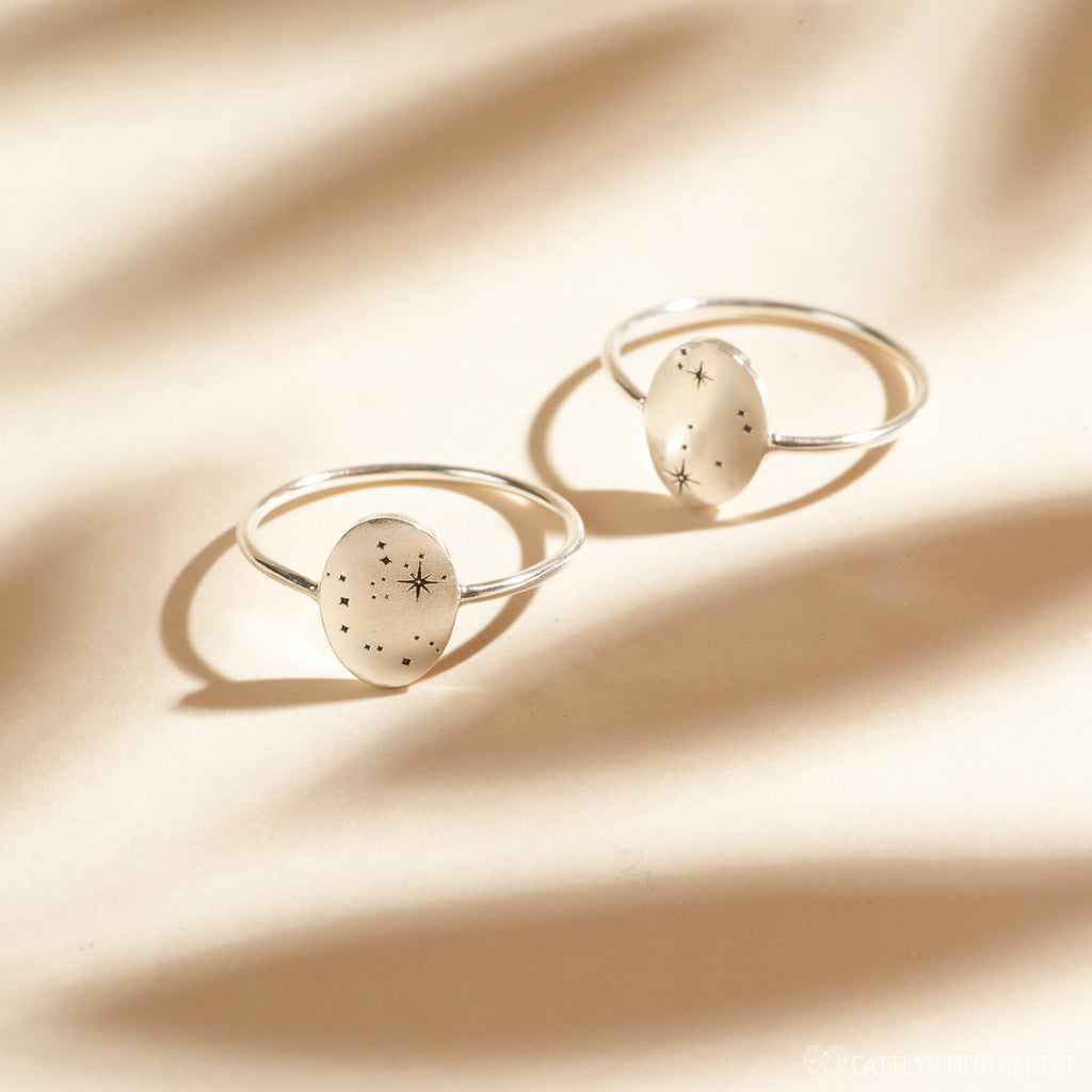 Two Constellation Oval Rings with silver bands and translucent oval stones featuring star and dot designs, displayed on a beige background.