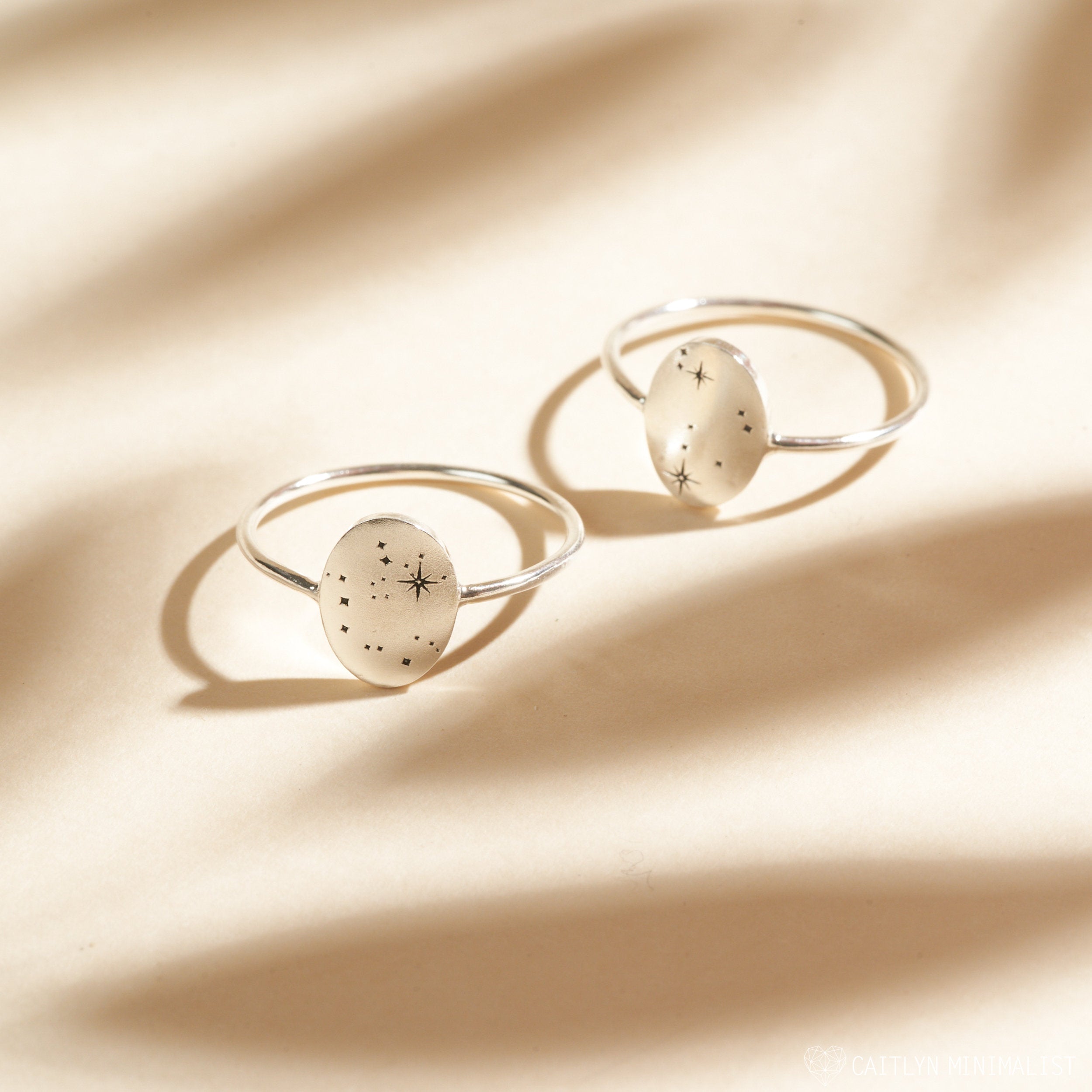 Two Constellation Oval Rings with silver bands and translucent oval stones featuring star and dot designs, displayed on a beige background.