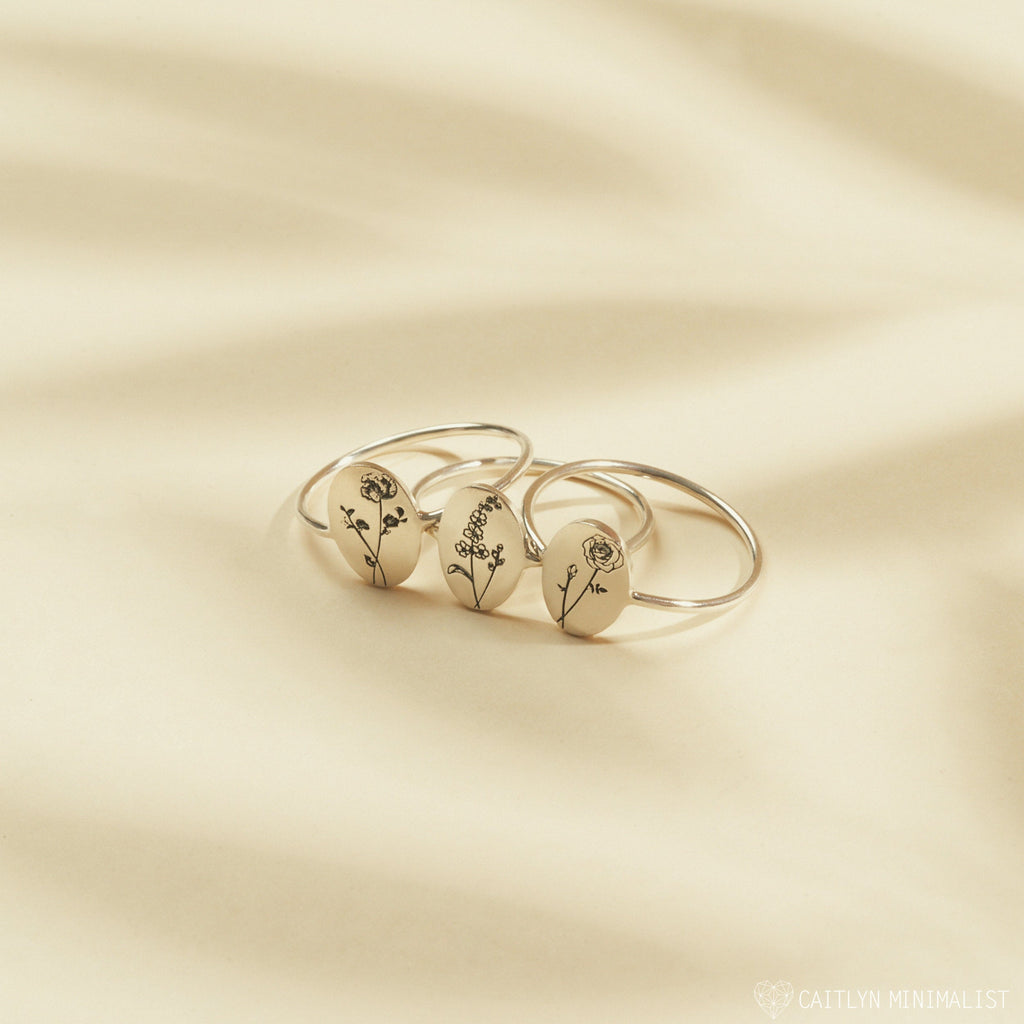 The Birth Flower Oval Ring, a silver signet ring engraved with flower designs for each birth month, is displayed on a light beige background.