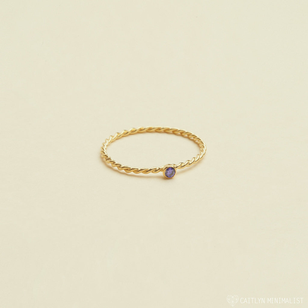 The Twisted Birthstone Ring features a delicate gold twist design with a small purple gemstone, perfect for adding color to your stack, displayed on a light beige background.