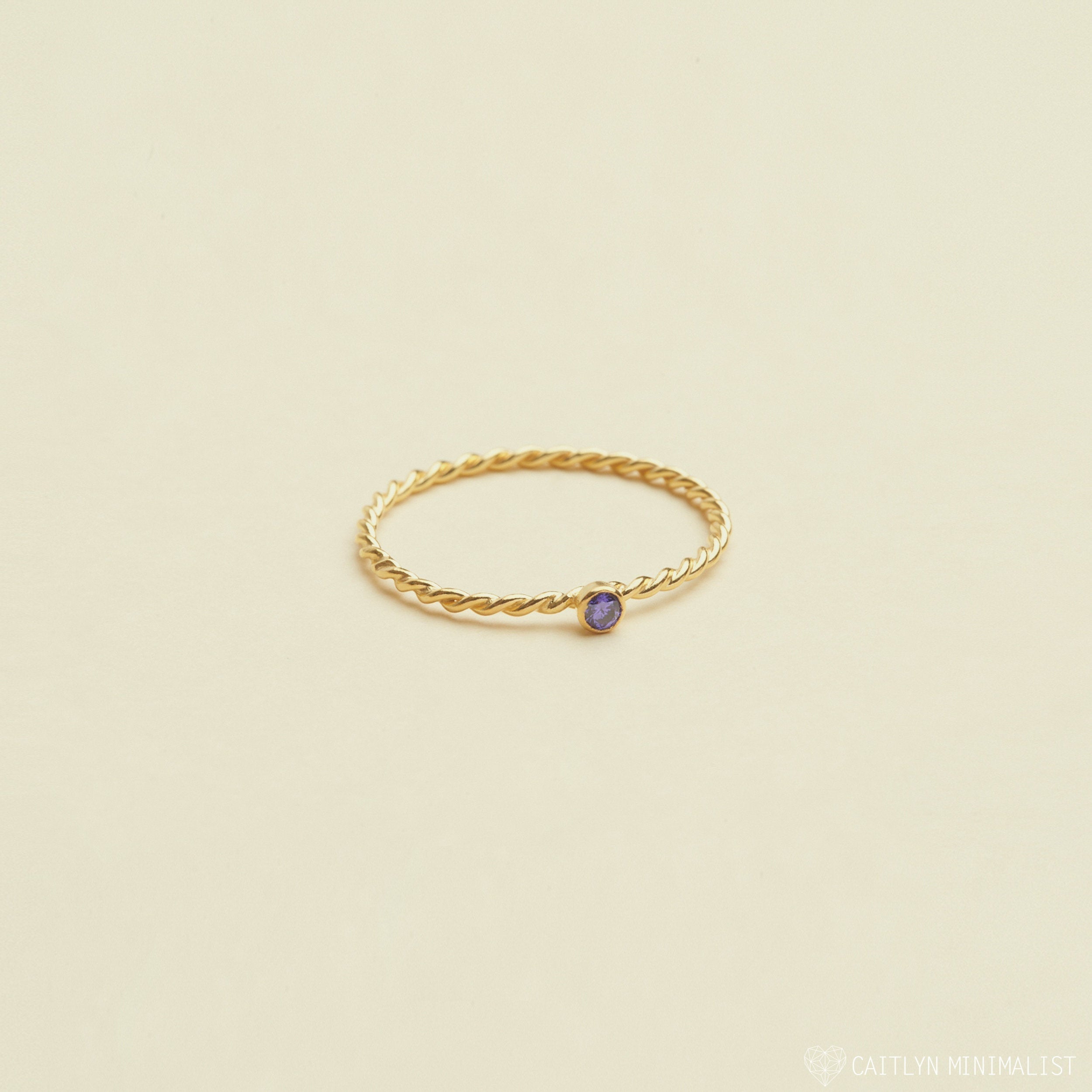 The Twisted Birthstone Ring features a delicate gold twist design with a small purple gemstone, perfect for adding color to your stack, displayed on a light beige background.