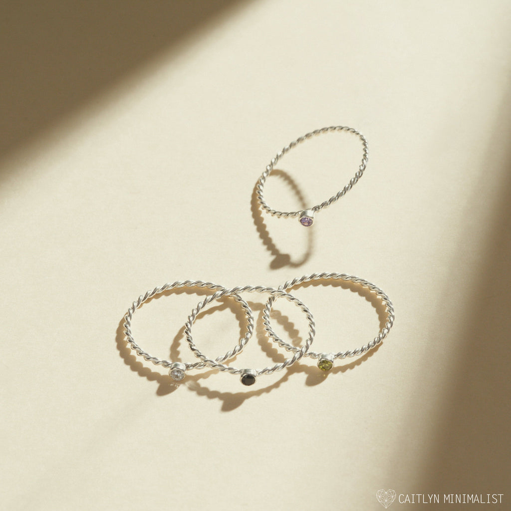 Four Twisted Birthstone Rings, each featuring a small gemstone, are elegantly stacked on a cream surface and illuminated by soft natural light.