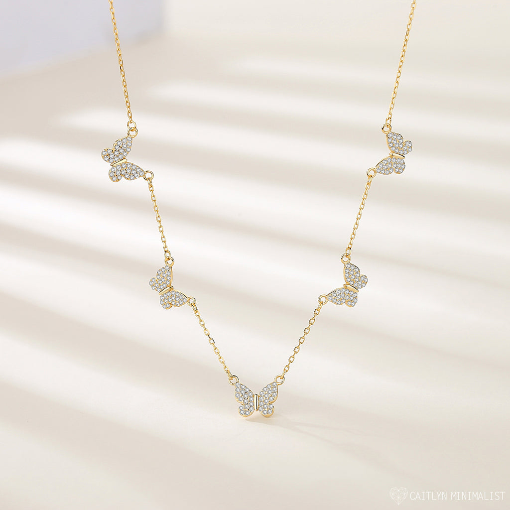 The Pave Butterfly Necklace features five diamond-encrusted butterfly pendants adorned with sparkling stones, elegantly displayed on a light background.