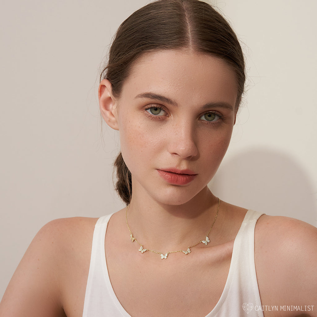 A woman with light skin and brown hair wears a white tank top and the Pave Butterfly Necklace, featuring diamond-encrusted butterflies for an elegant, feminine look.
