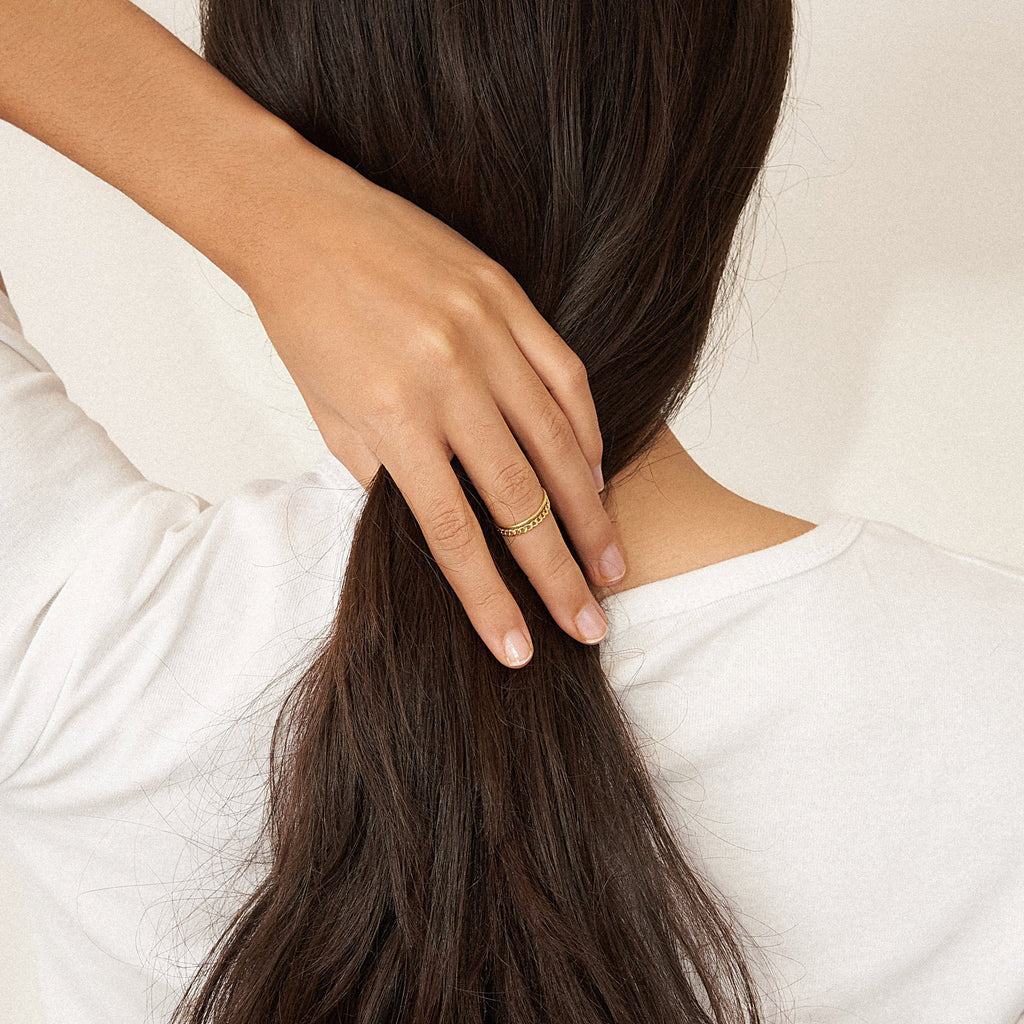 A person with long brown hair in a low ponytail, hand resting on head, wears a modern white shirt and a minimal gold ring from the Bold Stacking Ring Set.