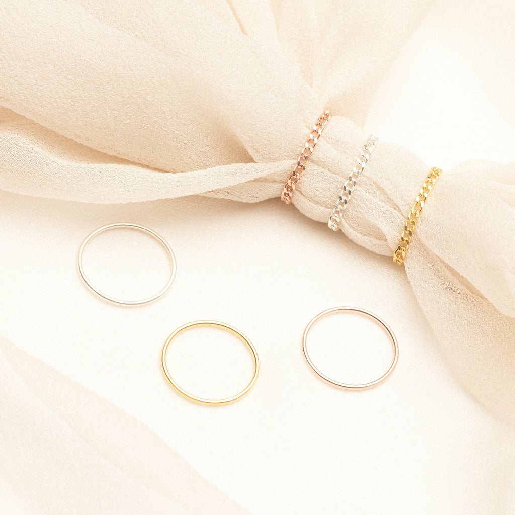 Three minimal rings from the Bold Stacking Ring Set in silver, gold, and rose gold are displayed on fabric and a cream background, adding a modern touch.