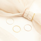 Three minimal rings from the Bold Stacking Ring Set in silver, gold, and rose gold are displayed on fabric and a cream background, adding a modern touch.