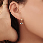 Close-up of a woman's ear wearing the Iris Huggies—gold huggie earrings with a small blue and white evil eye charm, perfect for adding stylish protection to your jewelry collection.
